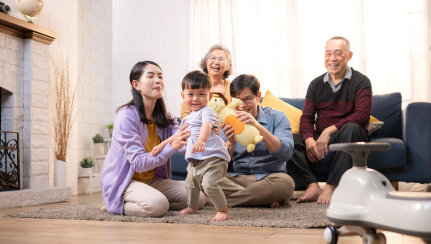 Happy multi-generational Asian family enjoying quality time together at home, cheering and smiling with a toddler riding a toy car in a cozy living room